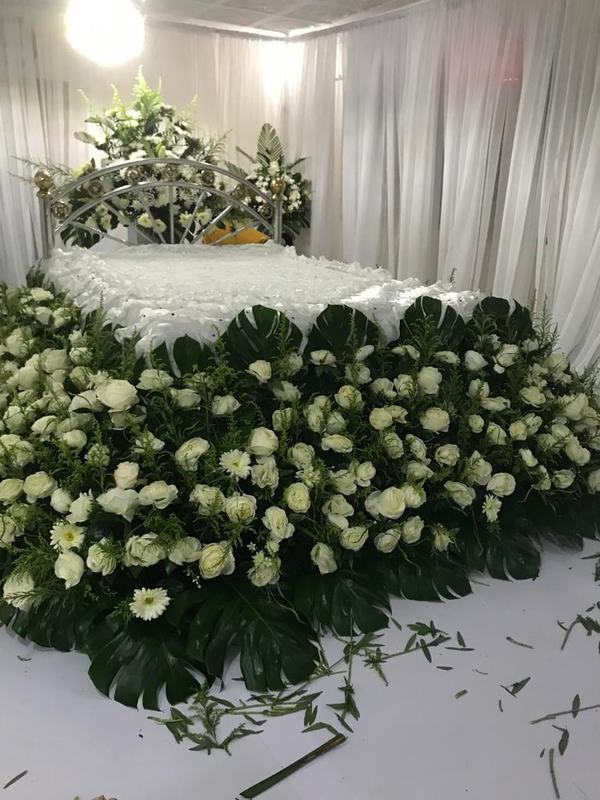 Large arrangement of white roses and green leaves around a draped memorial bed