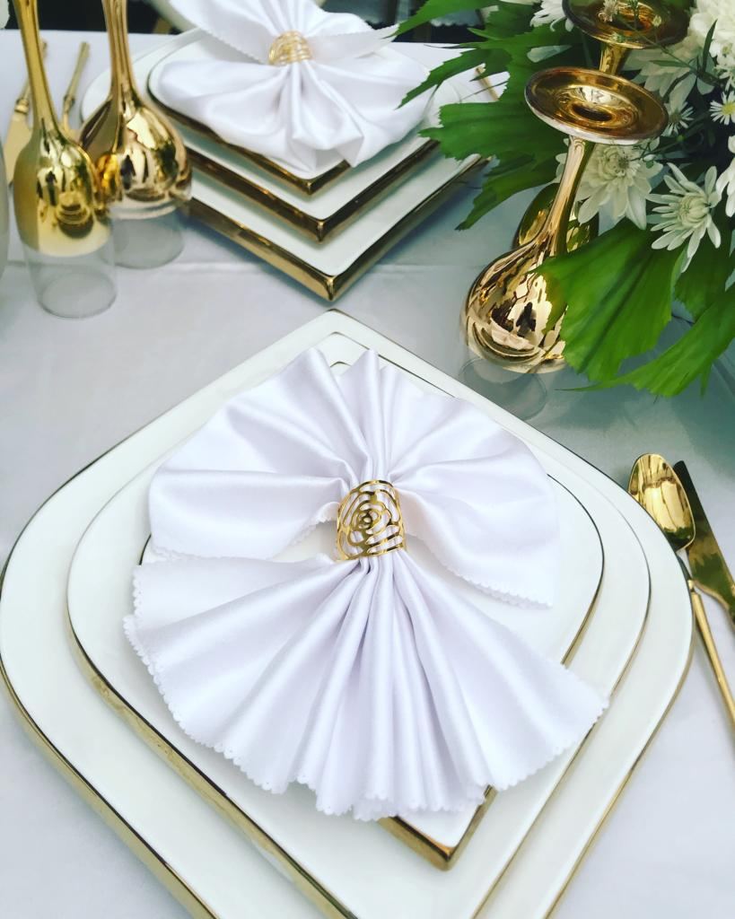 White napkin folded into a fan with a gold rose-shaped ring on a gold-rimmed white plate, beside gold candle holders and white flowers